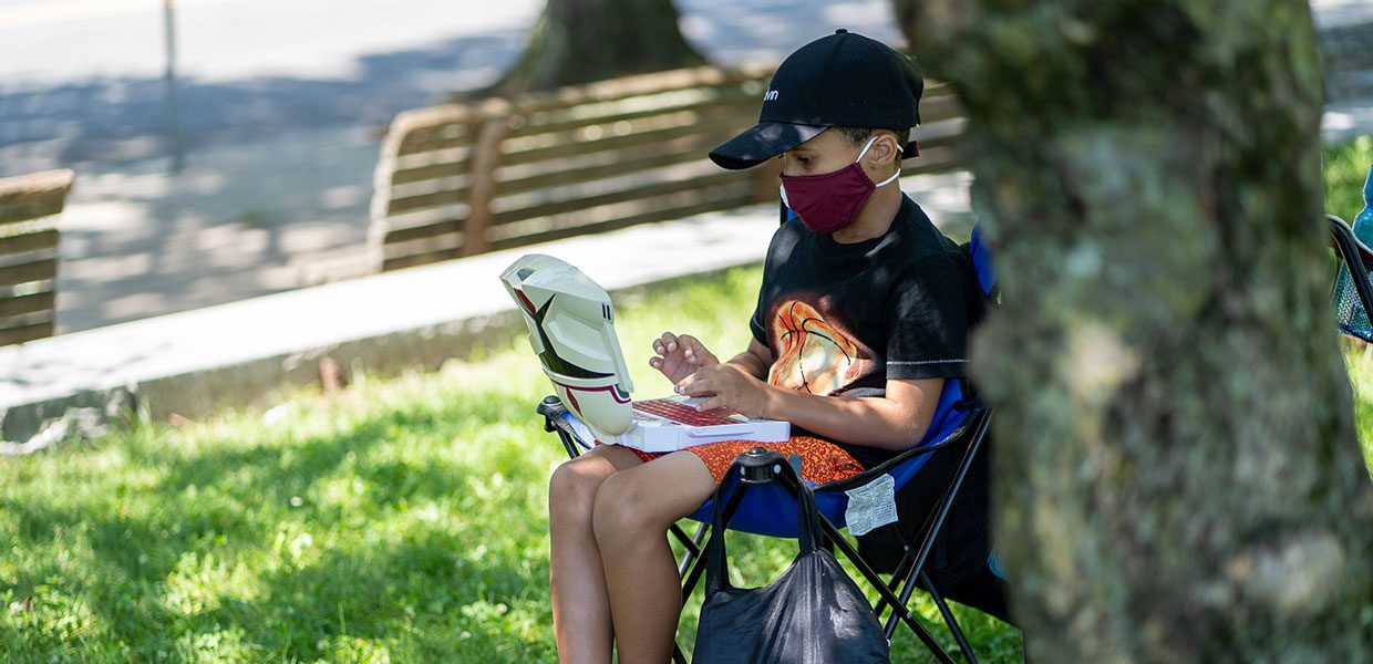 Kid with mask working on laptop under a tree outside. Photo credit: Sam Seidel