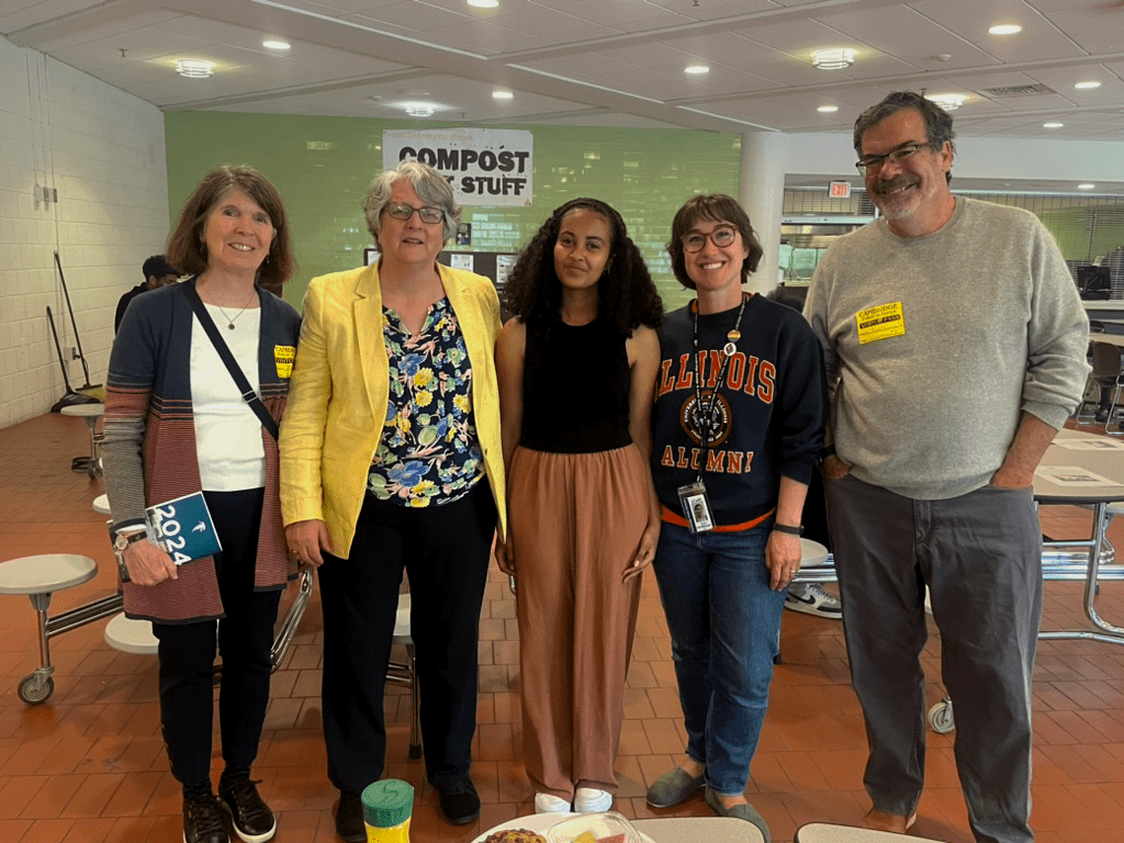 A group of five people standing together and smiling in a cafeteria setting. The group is diverse in age and attire, with a mixture of casual and professional clothing.